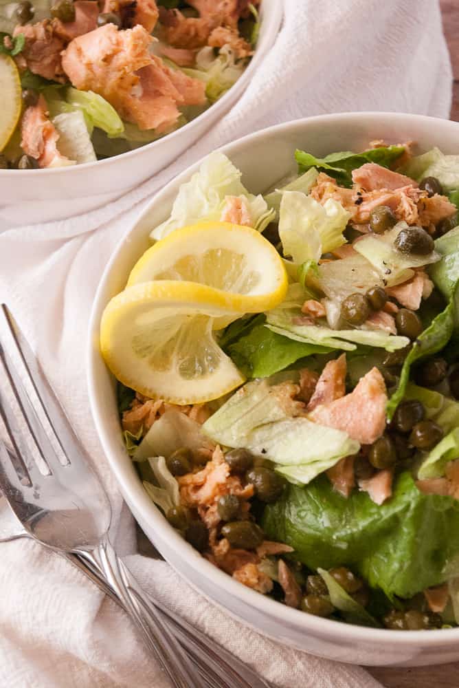 Salmon Piccata Salad in dinner bowls garnished with lemon slices.