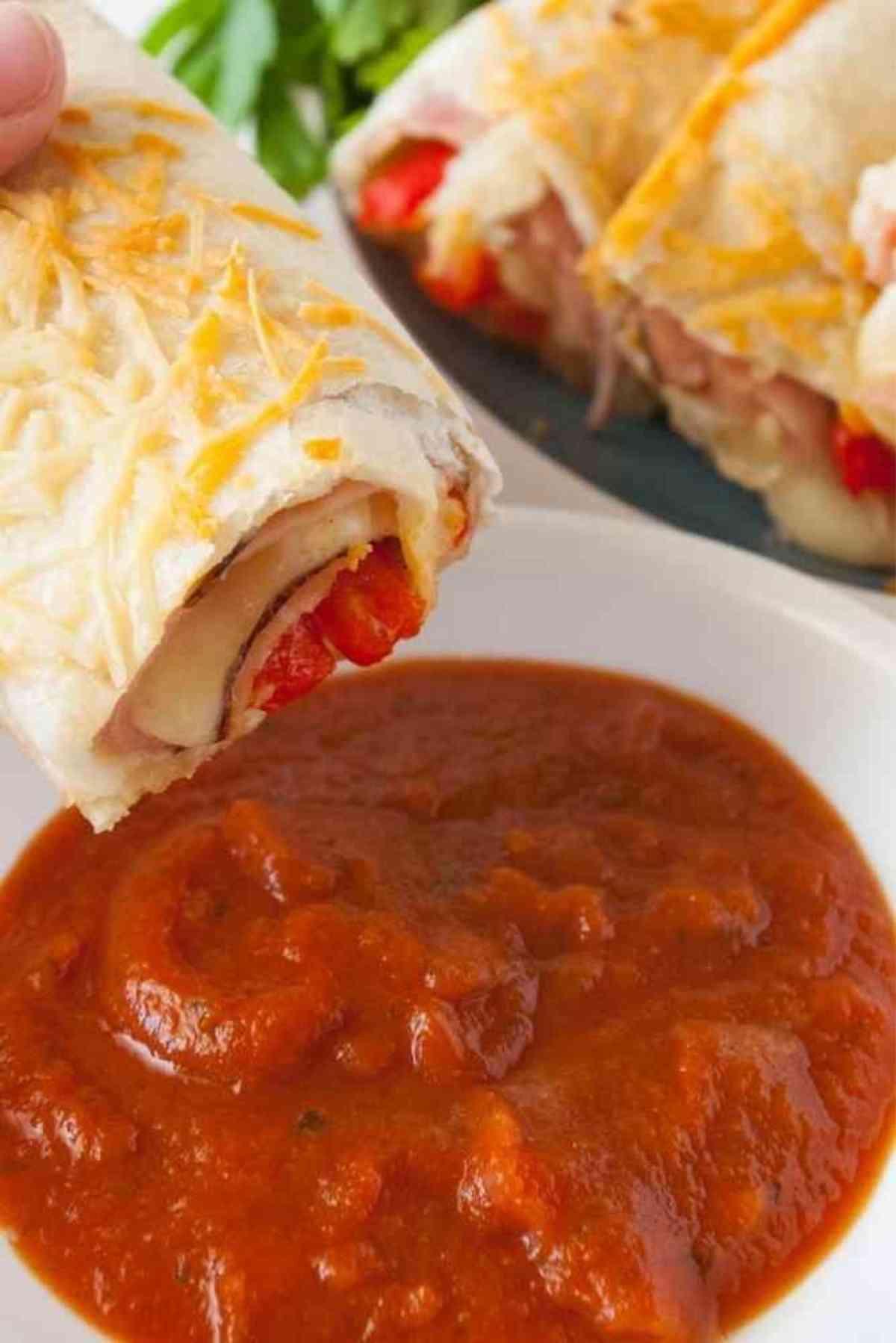 A hot ham and cheese roll up with a tortilla being dipped in pizza sauce.
