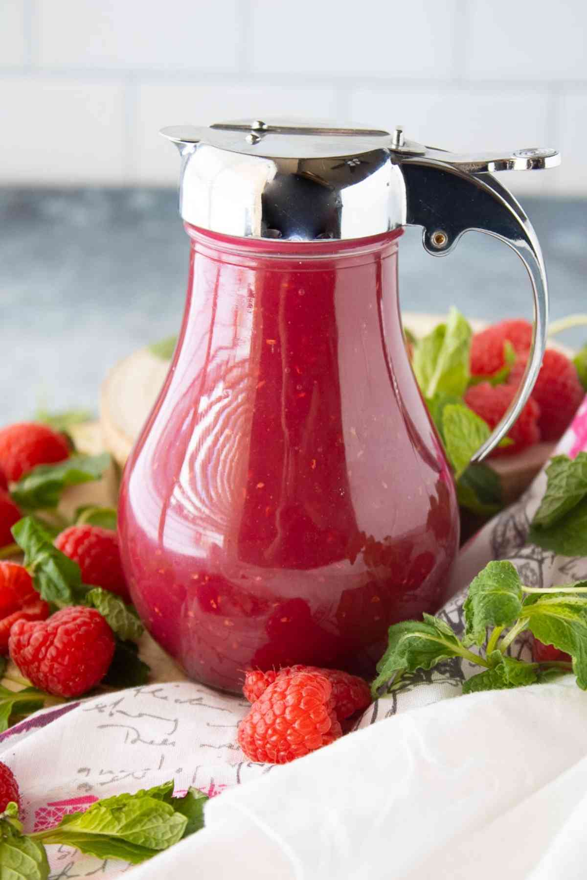 Bottle of raspberry cream syrup surrounded by fresh raspberries and mint.