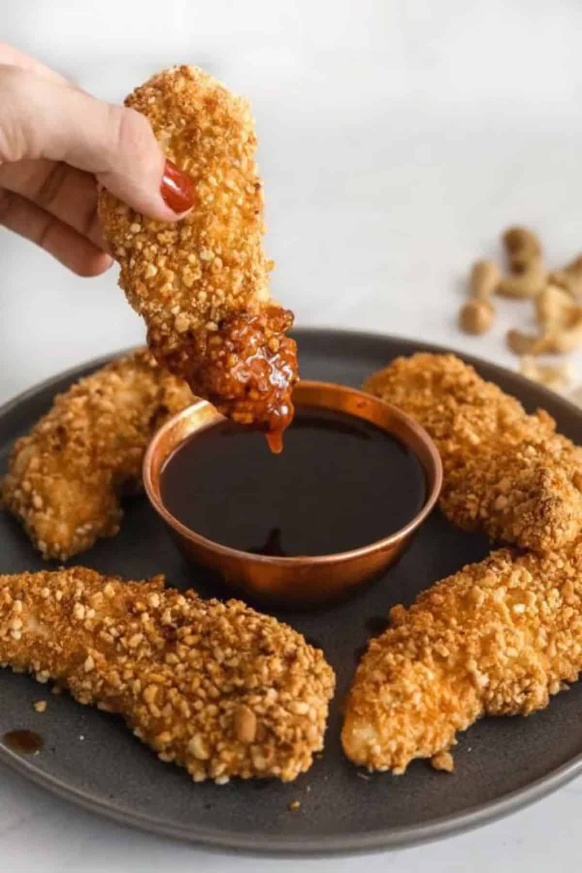 Baked Cashew-Crusted Chicken Fingers on a tray, one chicken finger dipped in a sauce.