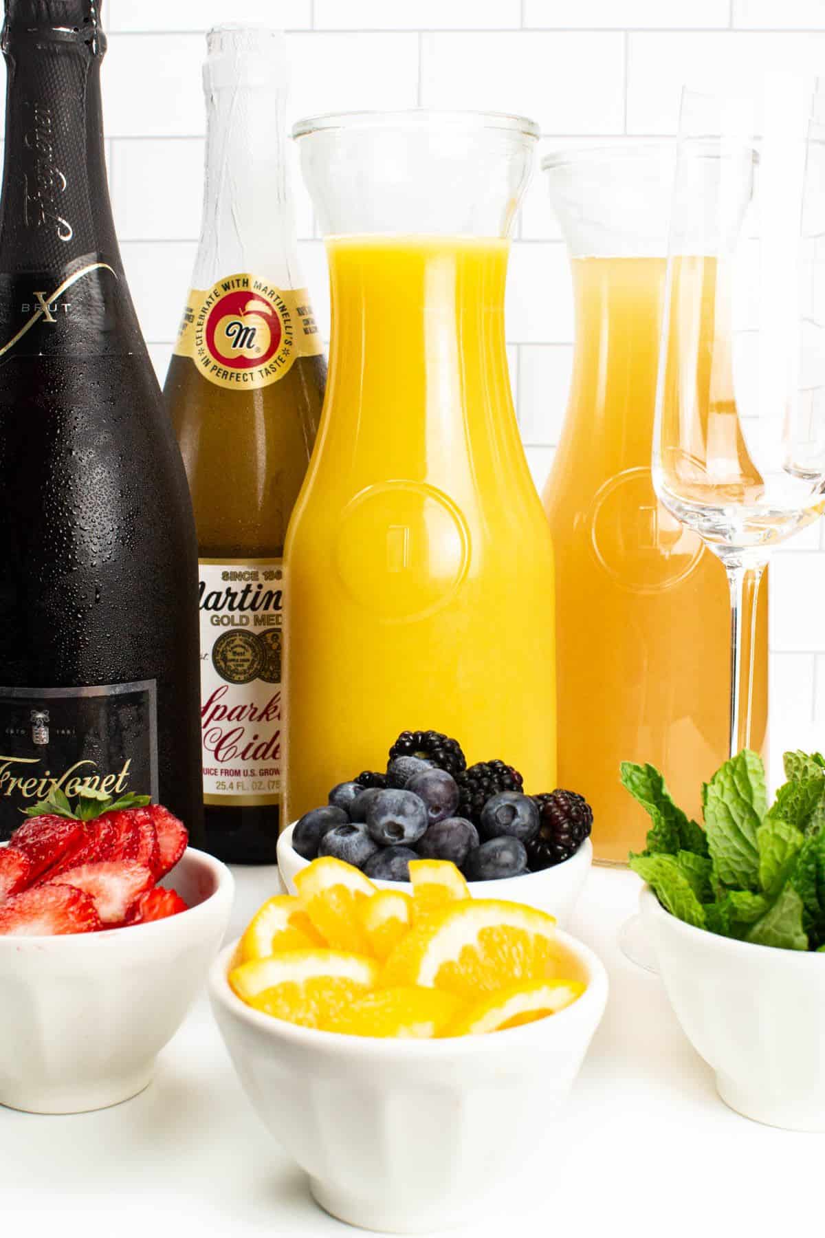 Different varieties of sparkling wines and juices in bottles, different varieties of fruits in white bowls.