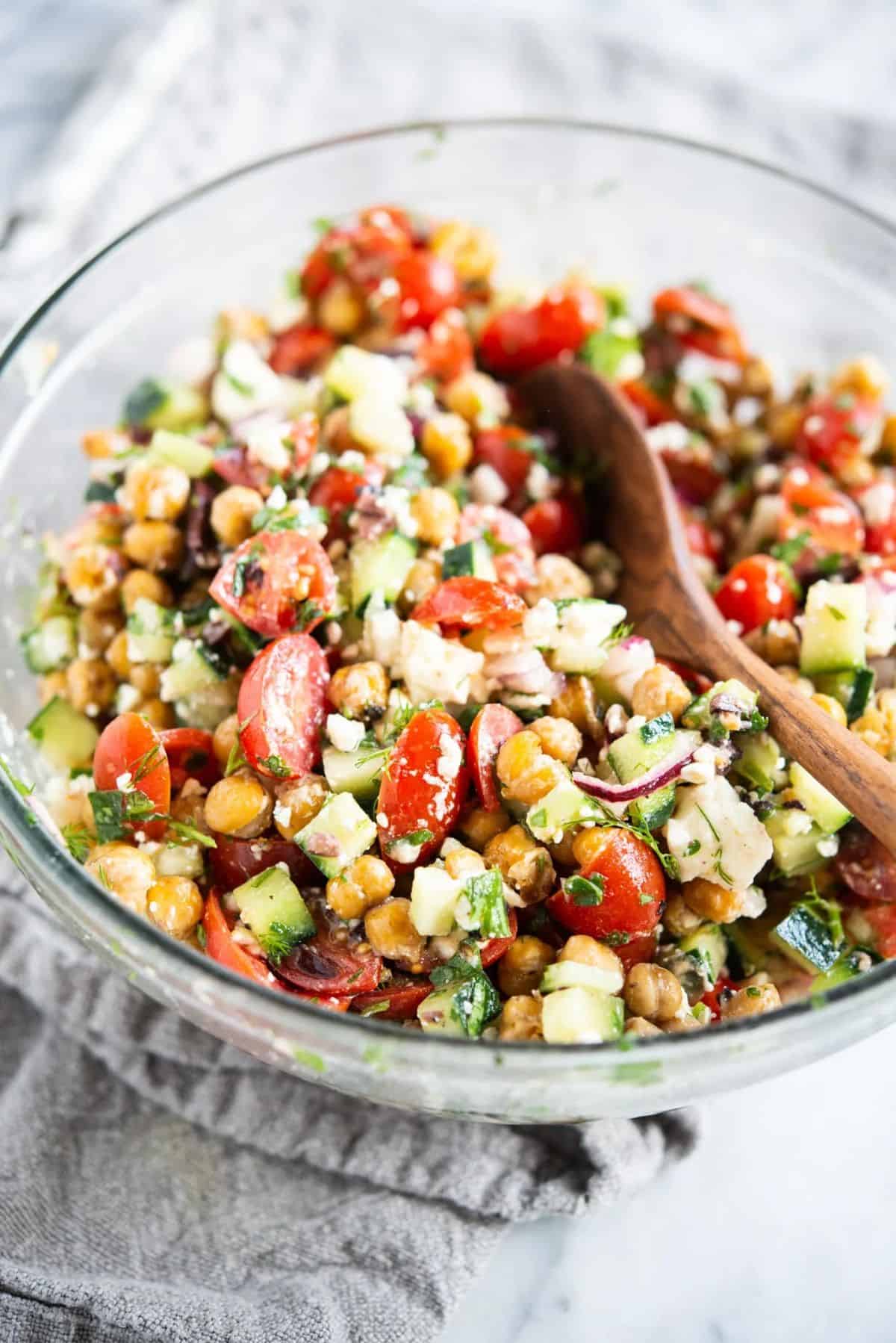 Healthy Mediterranean Chickpea Salad in a glass bowl with a wooden spatula.